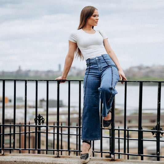 Woman in a white top and blue jeans standing on a railing with a cityscape in the background