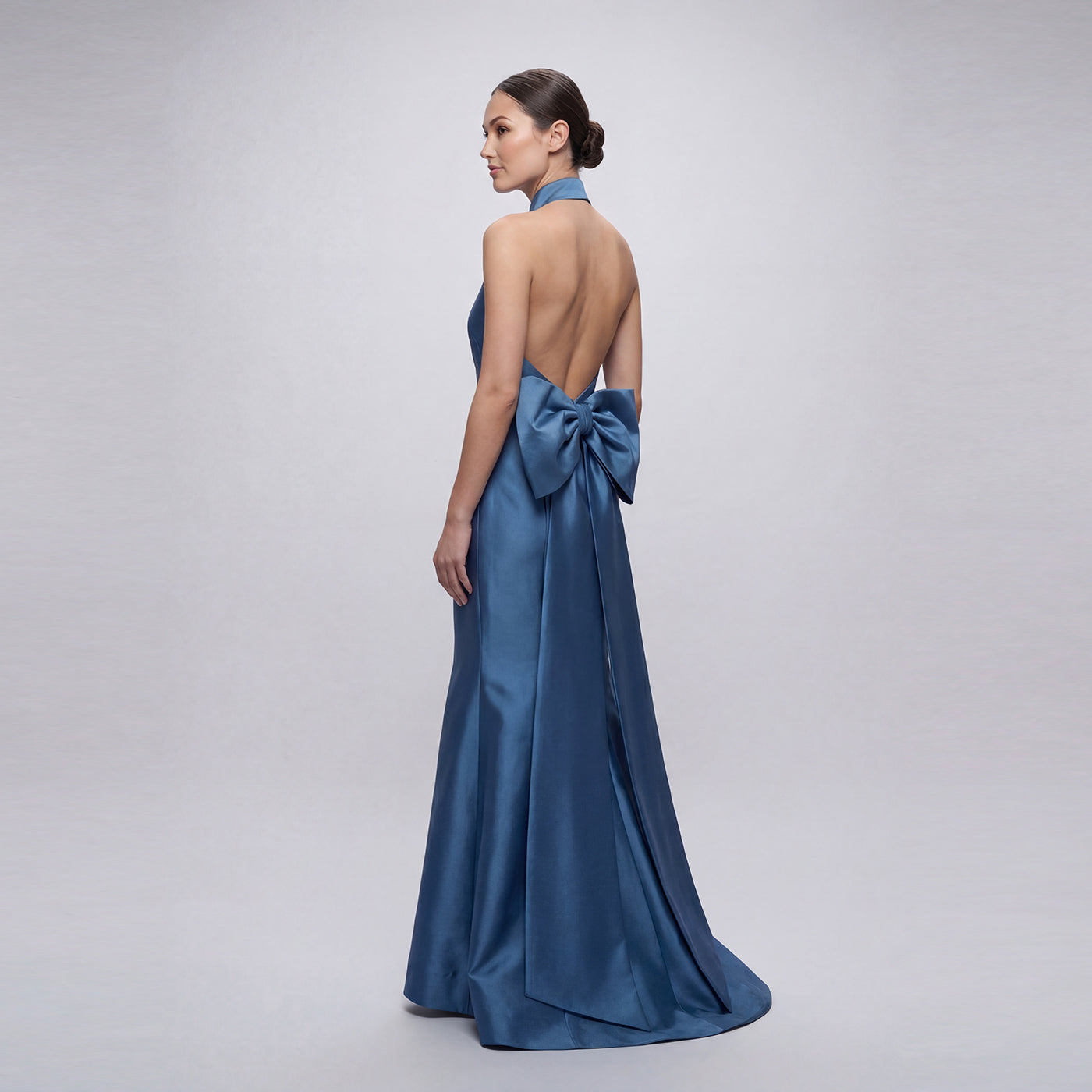 Blue evening gown with a large bow detail on the back, worn by a model against a gray background.