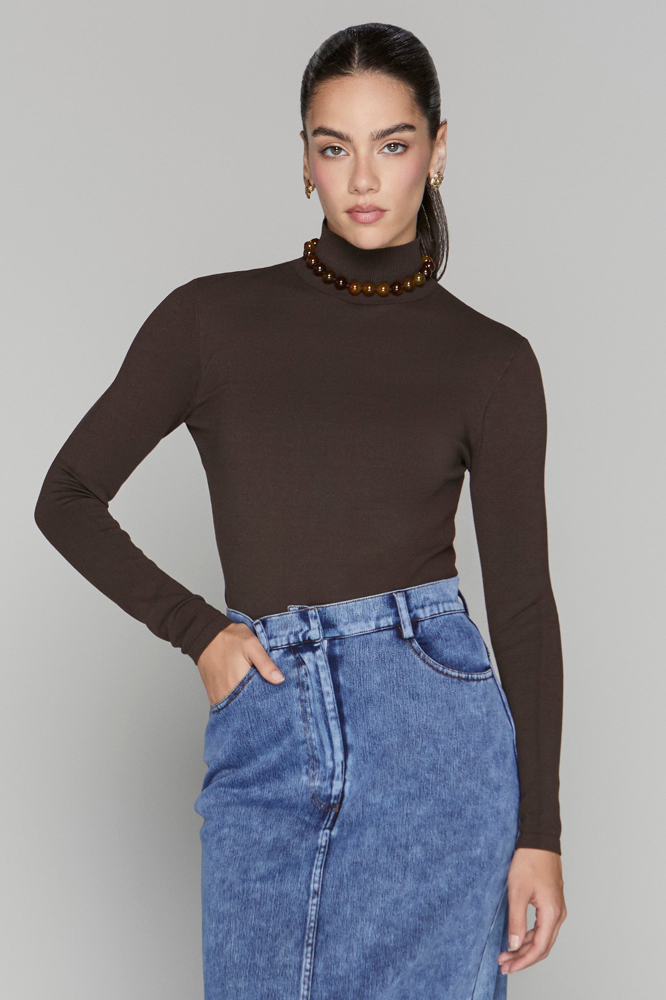 Woman wearing a brown turtleneck and blue jeans against a gray background