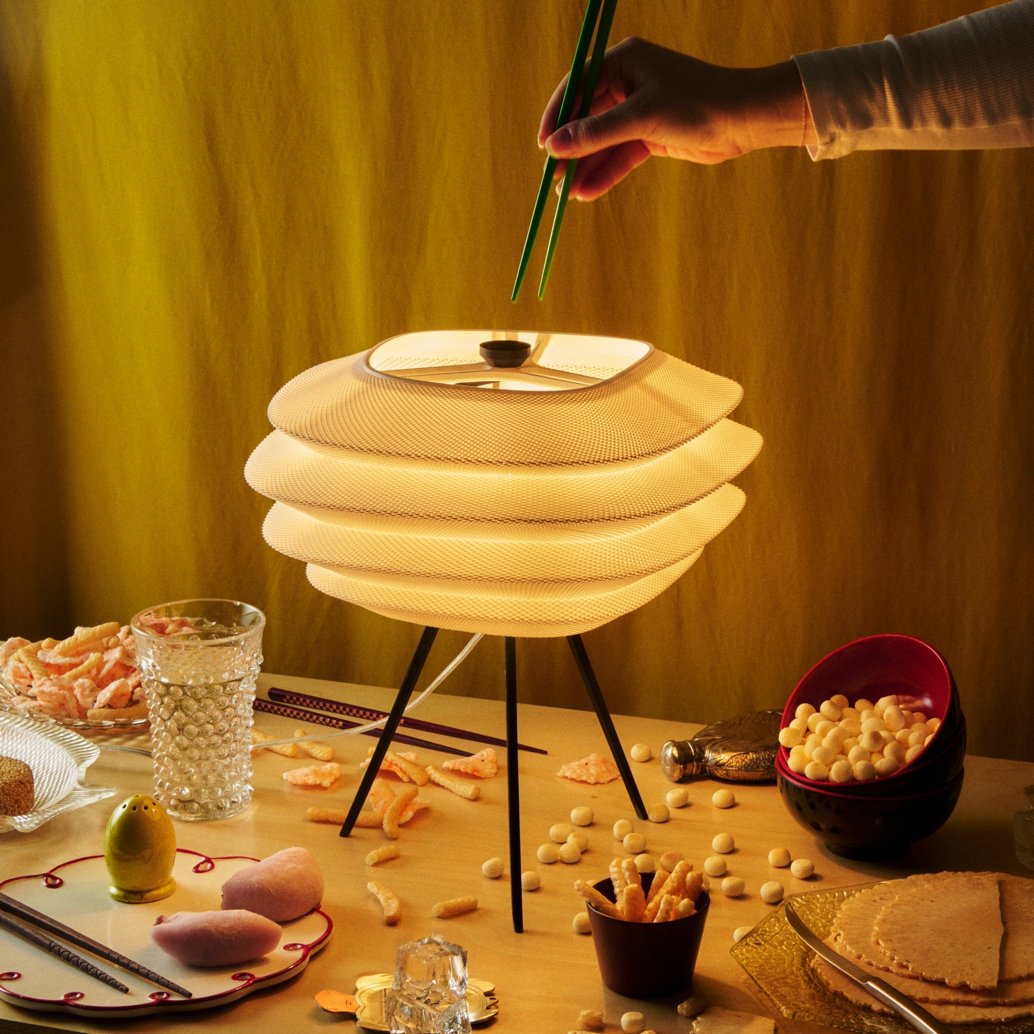 Person using chopsticks to pick up food from a table with a modern lamp and snacks.