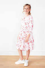 A young girl stands in a studio wearing a white three-quarter sleeve dress with a red bow and candy cane pattern, paired with white sneakers, against a plain white wall and light wood floor.