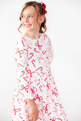 A young girl with brown hair in a red bow headband is wearing a white long-sleeve dress with a red and pink candy cane and bow pattern, photographed against a solid white background.
