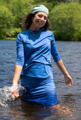 Woman in blue long-sleeve shirt and shorts standing in water with greenery in the background