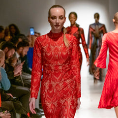 Model wearing a red patterned dress on a fashion runway with an audience.