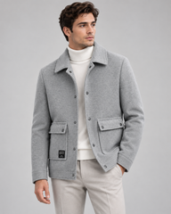 A man wearing a light gray wool-blend jacket over a white turtleneck sweater and light beige trousers, photographed against a neutral light gray studio background.