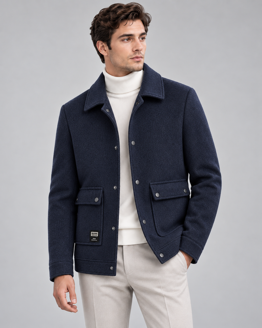 A man wearing a navy blue wool-blend jacket with silver snap buttons, a white turtleneck, and light beige trousers, photographed against a solid gray studio background.
