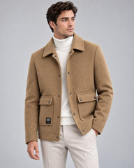 A man wearing a camel brown wool-blend jacket over a white turtleneck sweater and off-white trousers, standing against a solid gray studio background.