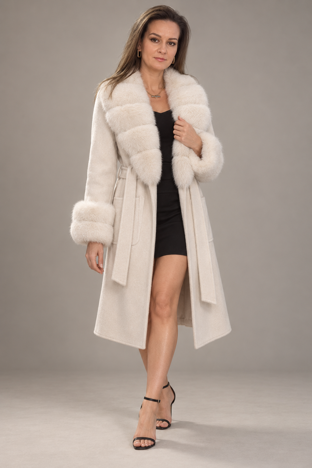 A woman modeling a long cream-colored faux fur coat with a matching belt, worn over a black dress and black high-heeled sandals, against a neutral gray studio background.