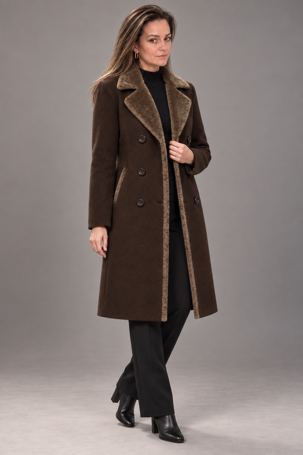 A woman models a long brown double-breasted coat with a brown faux-shearling collar and lining, styled with a black turtleneck, black trousers, and black ankle boots against a solid gray studio background.