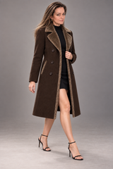 A woman models a long brown suede-like coat with a contrasting brown faux-fur collar and lining, worn over a black dress and black high-heeled sandals against a neutral gray studio background.