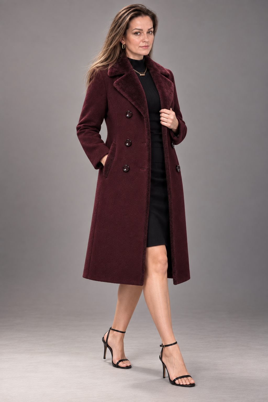 A woman modeling a long double-breasted coat in deep burgundy over a black dress and black strappy heels, photographed against a neutral gray studio background.