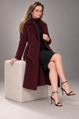 A woman wearing a long burgundy wool-blend coat over a black dress and black high-heeled sandals, seated on a light gray cube against a neutral gray studio background.
