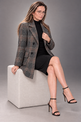 A woman wearing a black dress and black high-heeled sandals sits on a light gray upholstered cube, wearing a patterned double-breasted blazer over a black top. The blazer features a subtle plaid/check pattern in black, gray, and brown tones.