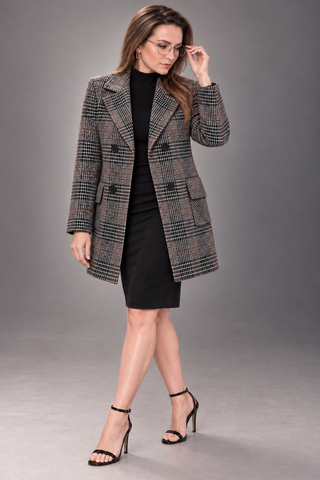 A woman models a double-breasted plaid blazer over a black dress and black high-heeled sandals against a solid gray studio background.