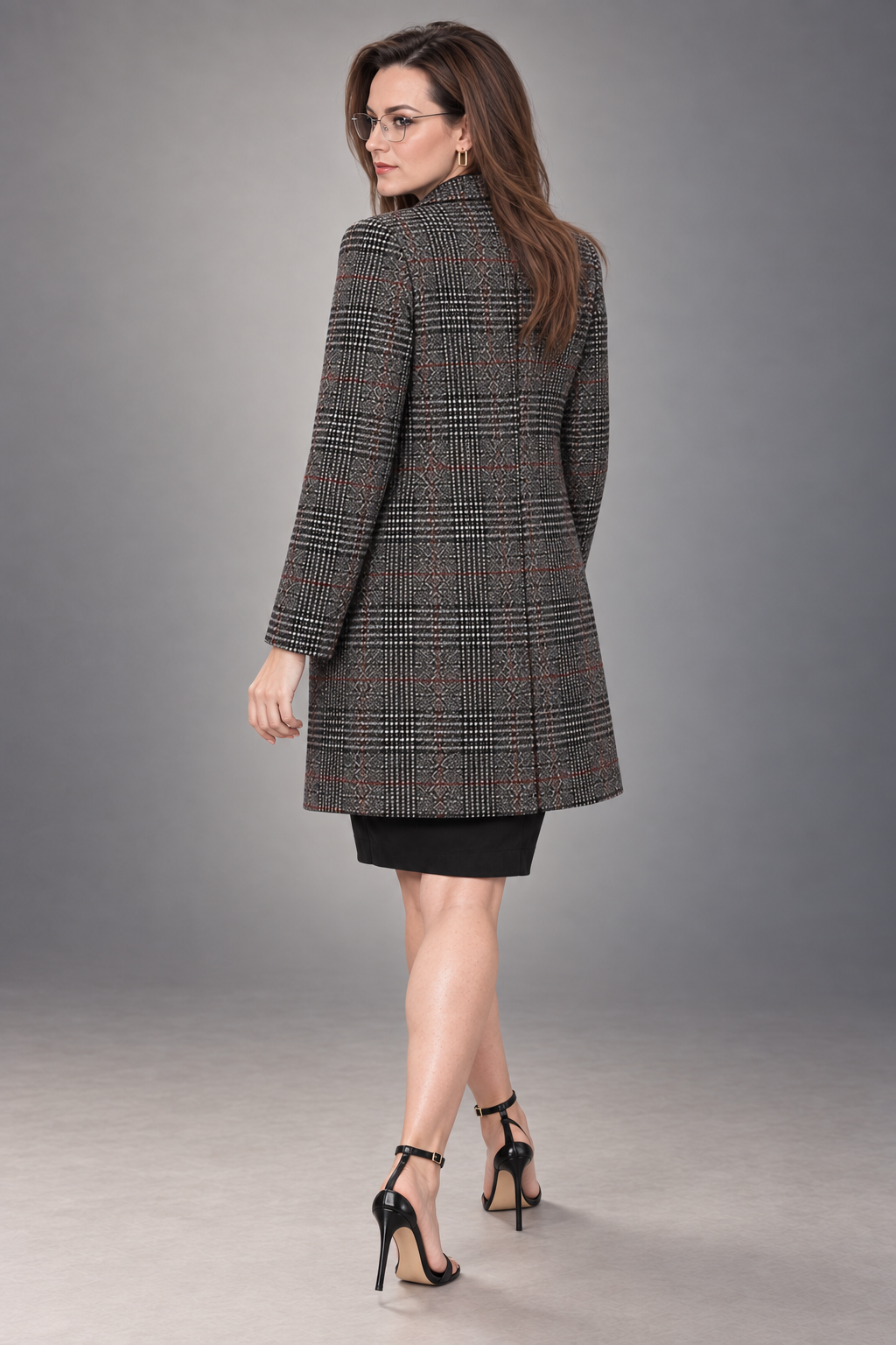 A woman modeling a mid-length black, gray, and rust-red patterned blazer from the back, paired with a black skirt and black high-heeled sandals, against a neutral gray studio background.