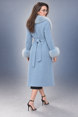 A woman modeling a light blue mid-length coat with a matching waist tie and fluffy light blue fur trim at the collar and cuffs, photographed against a smooth gray studio background.
