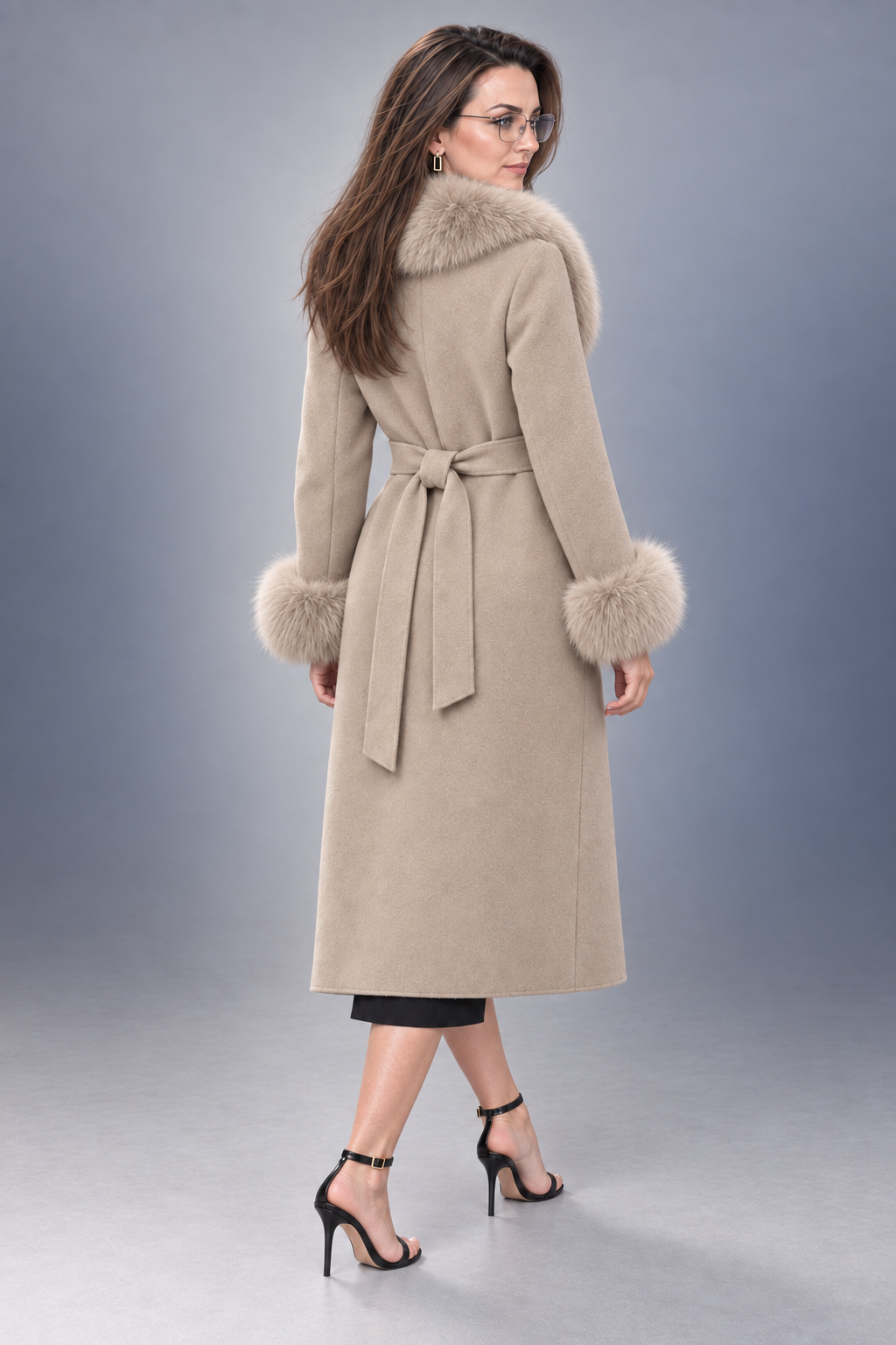 A woman modeling a mid-length beige coat with a plush beige fur collar, matching fur cuffs, and a self-tie belt, shown from a three-quarters back view against a gray studio background.
