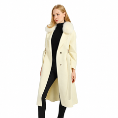 A woman modeling a long cream-colored coat with a fluffy white fur-trimmed collar, worn over a black turtleneck and black leggings, against a solid white background.