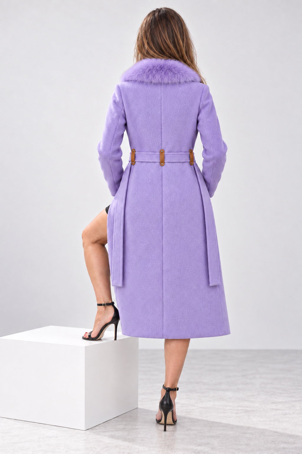 A woman shown from the back wearing a lavender purple mid-length coat with a fuzzy collar and a brown belt, standing on a white display block in a studio setting.