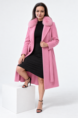 A woman models a long pink coat with a fluffy faux-fur collar, worn over a black dress and black heeled sandals, standing on a white block against a light gray studio background.
