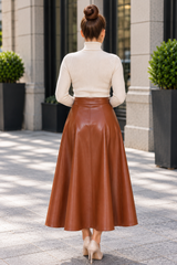 A woman viewed from behind wearing a cream mock-neck long-sleeve top and a high-waisted, flared brown faux-leather midi skirt, standing on a paved outdoor walkway.