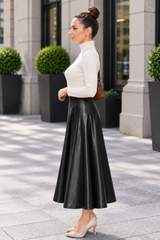 A woman stands outdoors in a city setting wearing a fitted white mock-neck long-sleeve top and a high-waisted black faux-leather A-line midi skirt, paired with beige heels and a brown shoulder bag.