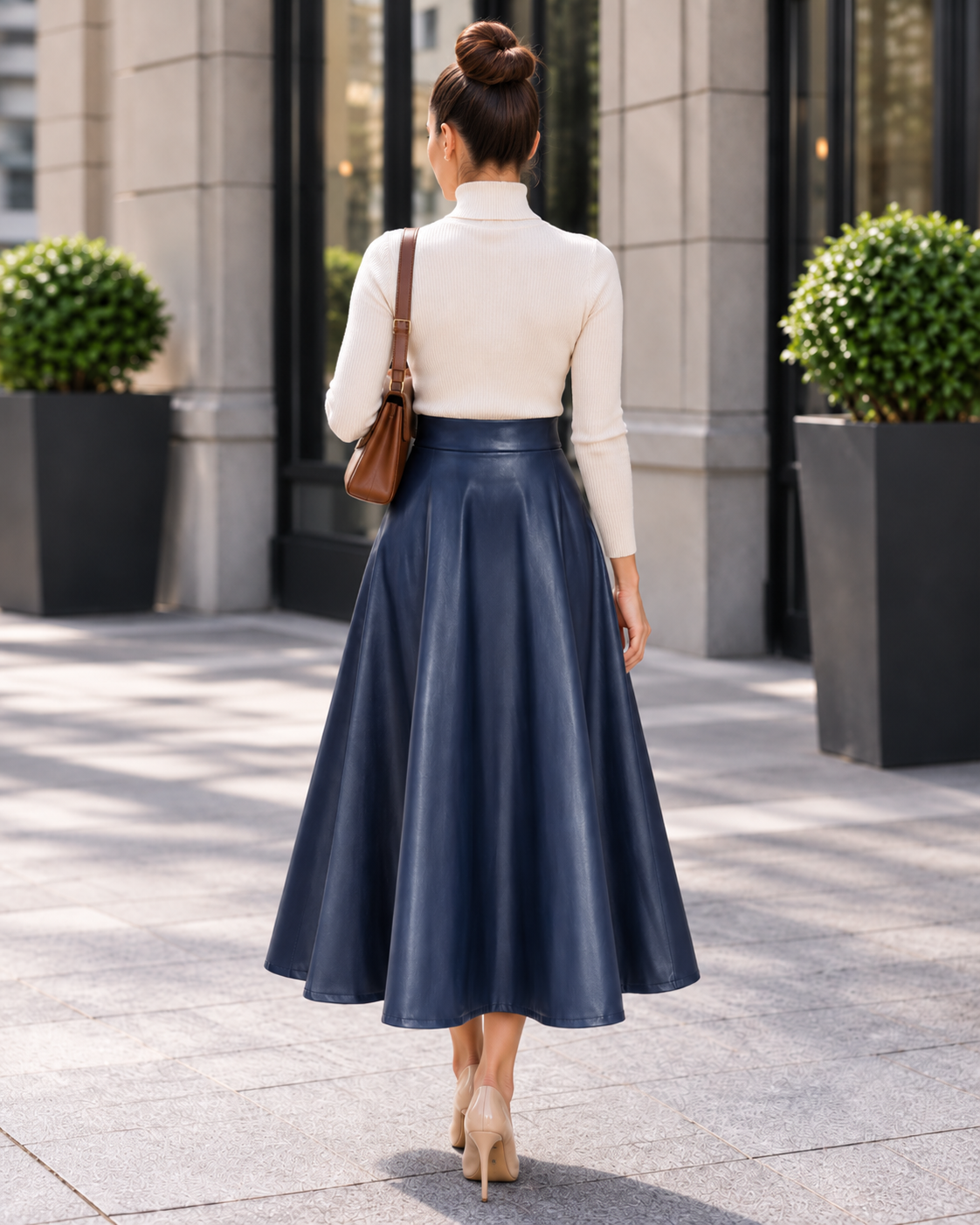 A woman viewed from behind wearing a cream mock-neck long-sleeve top and a navy blue faux-leather midi skirt, carrying a brown shoulder bag while walking on a paved outdoor walkway.