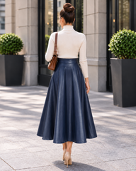 A woman viewed from behind wearing a cream mock-neck long-sleeve top and a navy blue faux-leather midi skirt, carrying a brown shoulder bag while walking on a paved outdoor walkway.