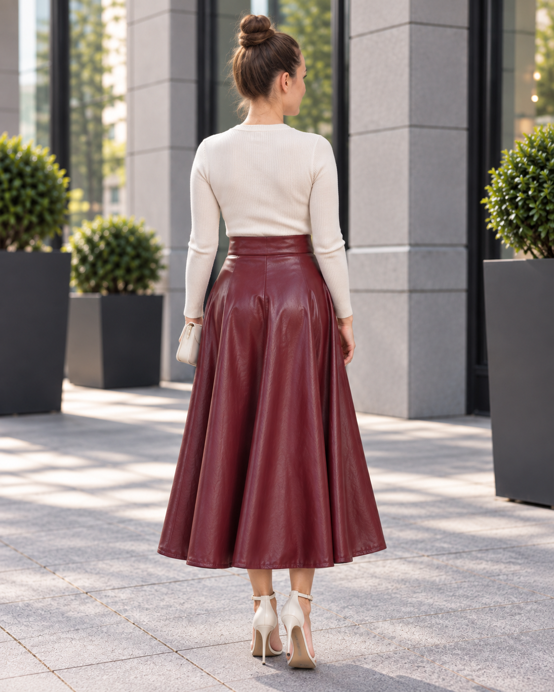 A woman viewed from behind wearing a cream long-sleeve knit top and a high-waisted, flared burgundy faux-leather midi skirt, standing on a paved outdoor walkway.