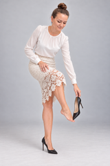 A woman modeling a white long-sleeve blouse and a cream lace pencil skirt, holding a black high heel shoe against a light gray studio background.