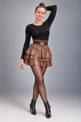 A woman modeling a black long-sleeve top, a brown tiered faux-leather mini skirt with a black belt, sheer black tights, and black ankle boots against a gray studio background.
