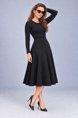 A woman modeling a black long-sleeve midi dress with a flared skirt, paired with black pointed-toe heels and sunglasses, against a light gray studio background.