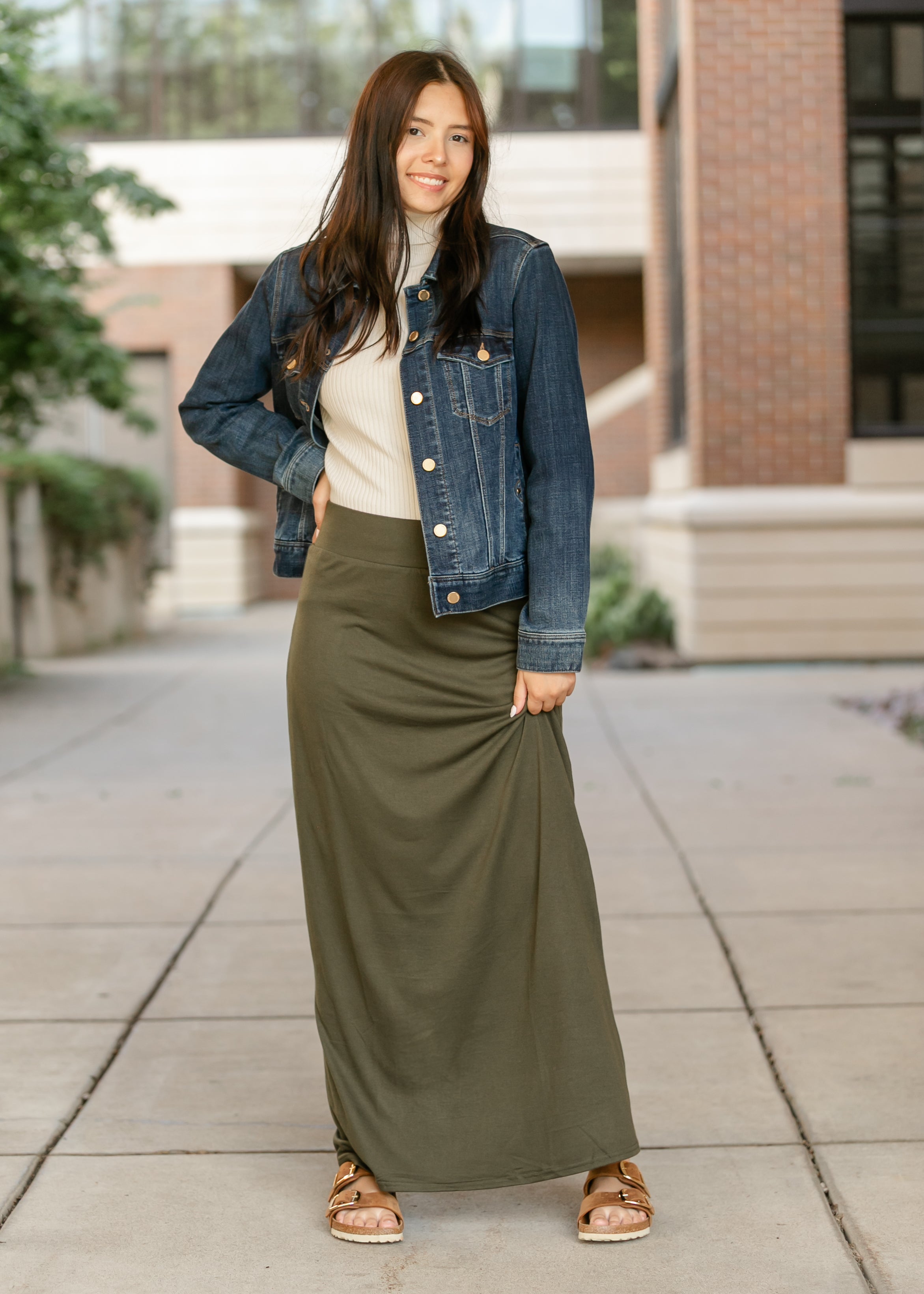 Woman wearing a denim jacket and long green skirt standing on a sidewalk.
