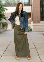 Woman wearing a denim jacket and long green skirt standing on a sidewalk.