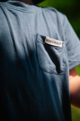 Blue t-shirt with a brand label on the chest pocket against a blurred green background
