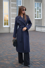 Woman wearing a long navy coat with a belt, standing on a brick pavement.