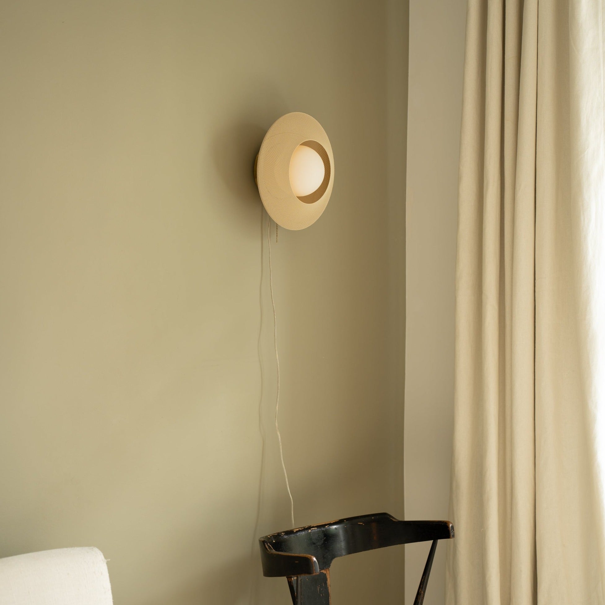 Round wall light fixture on a beige wall with a curtain to the right.