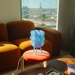 Blue lamp on a red stool in front of an orange sofa with a cityscape view through a window.
