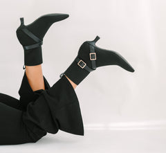 Black high-heeled boots with gold buckles worn by a person in a black outfit on a light gray background