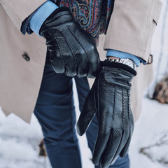 A close-up of a person wearing black leather gloves, a beige coat, a blue shirt, and a patterned scarf, standing outdoors in snow.