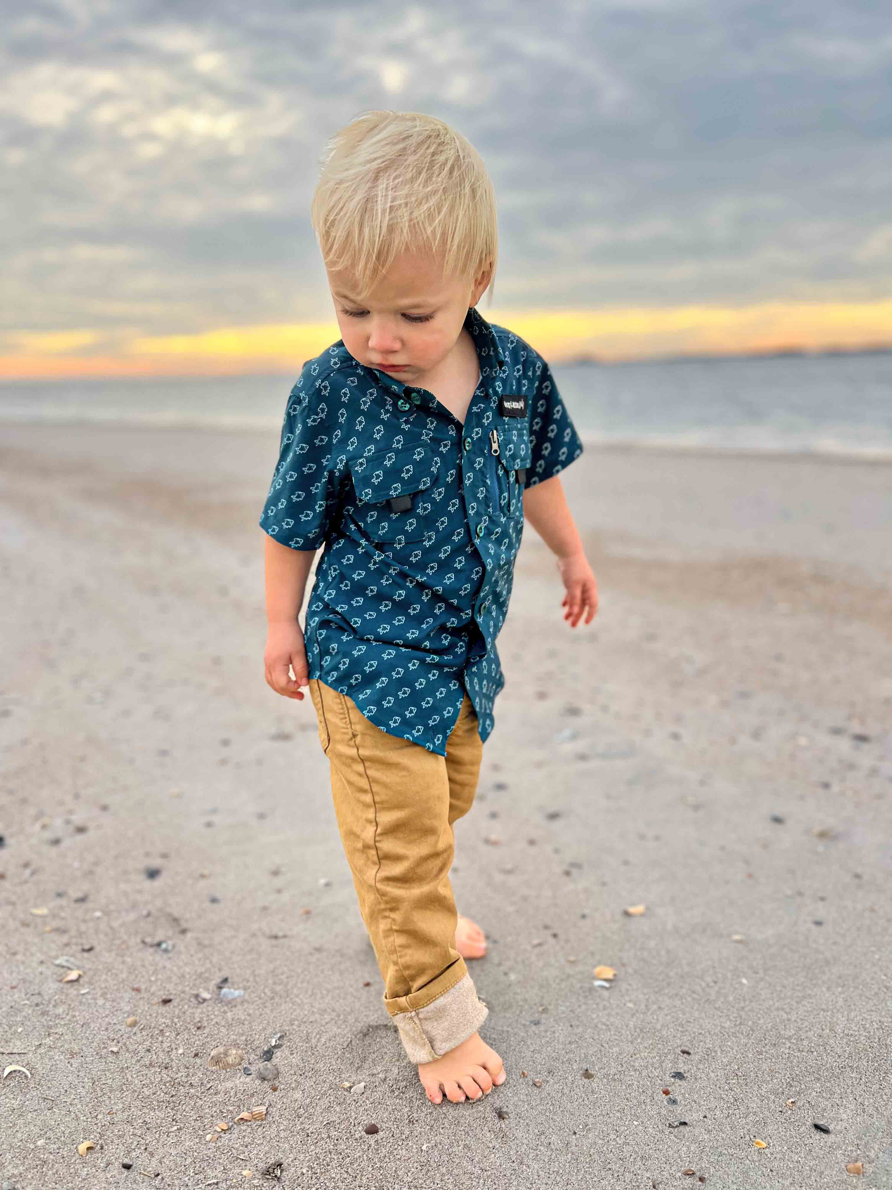 Child in a blue shirt and beige pants standing on a beach with a sunset in the background