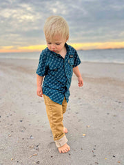 Child in a blue shirt and beige pants standing on a beach with a sunset in the background