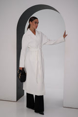 Woman wearing a white coat and black pants, holding a black handbag, against a plain background.