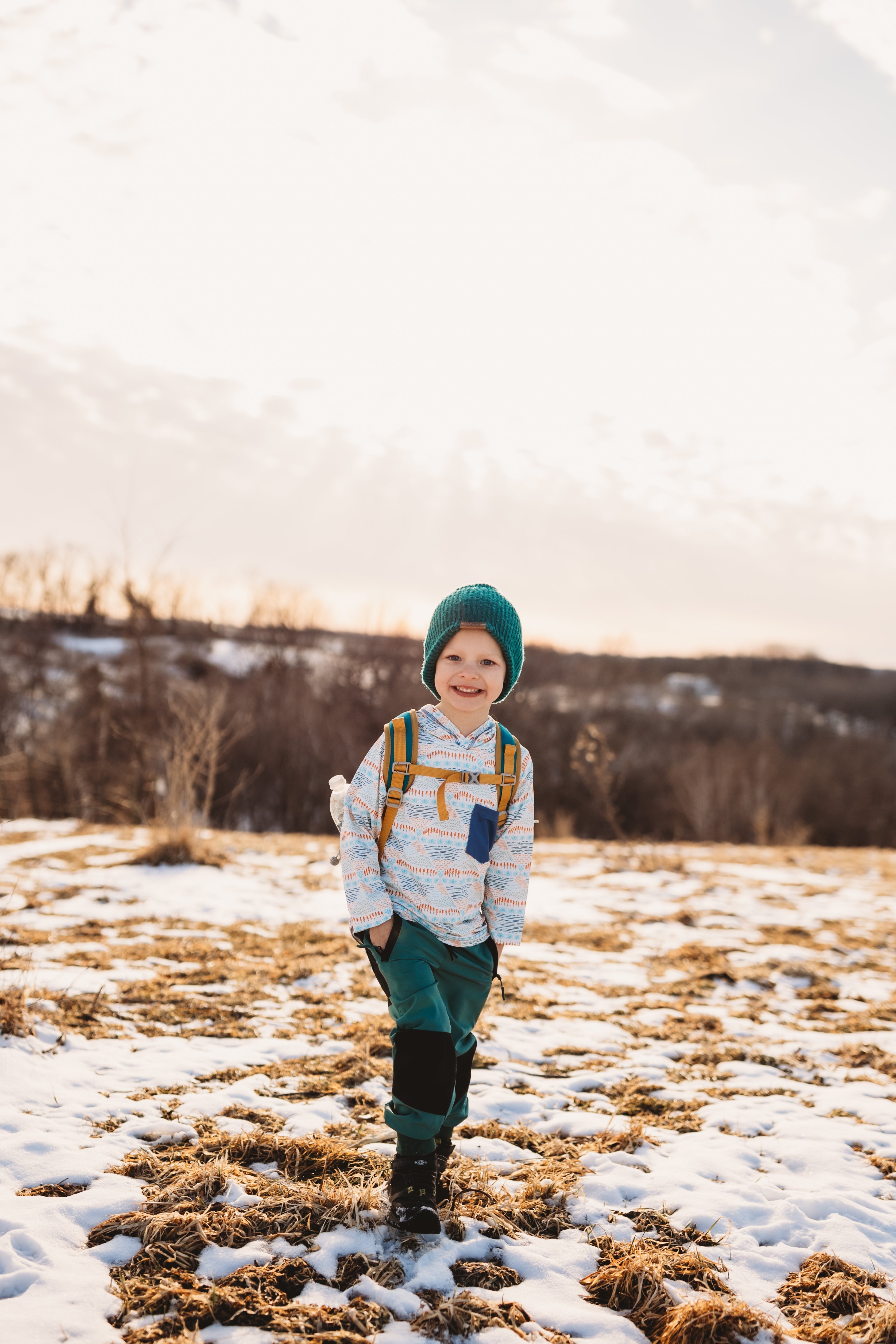 Child in a snowy field wearing a green hat and backpack
