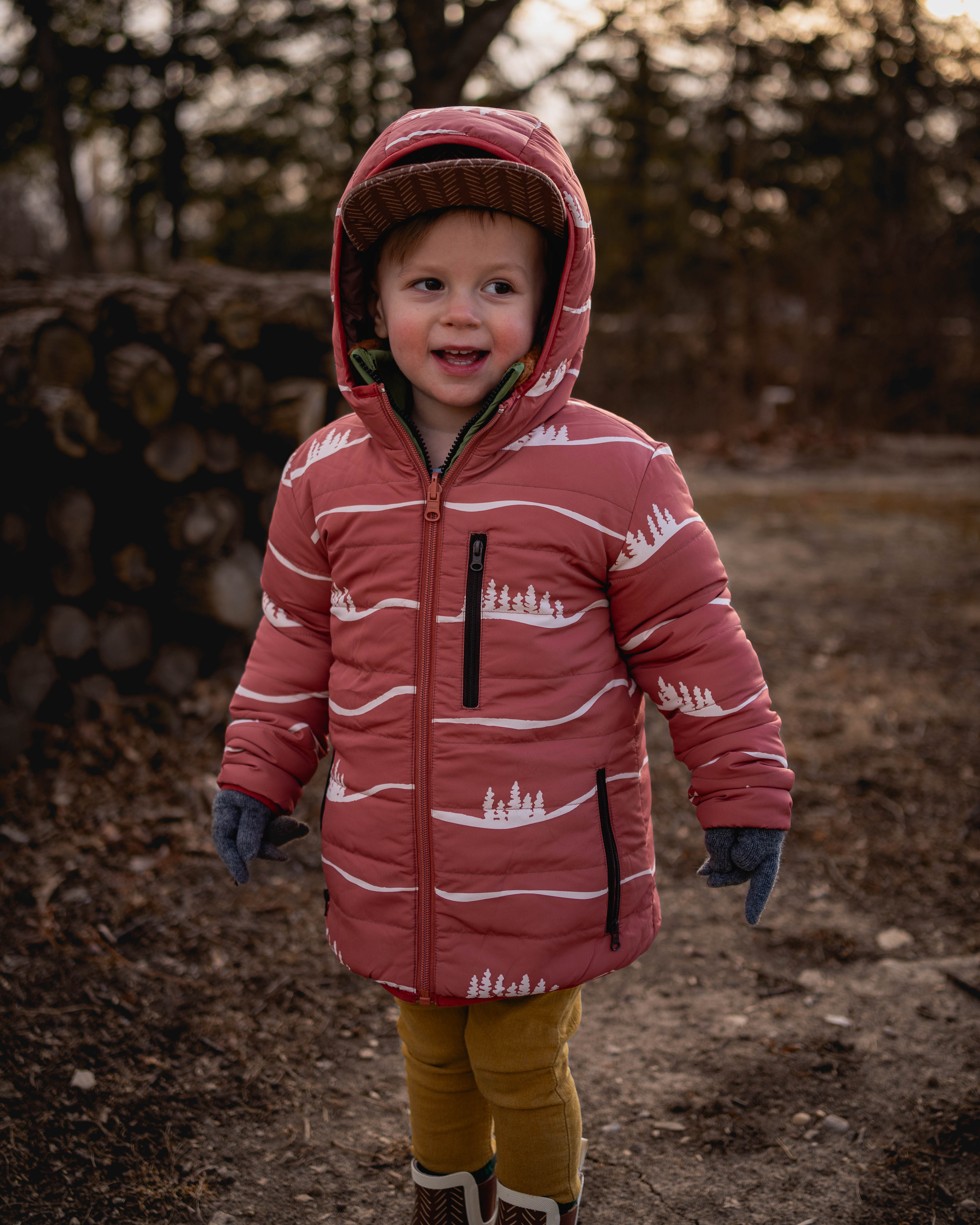Child wearing a red jacket with white patterns outdoors near stacked logs.