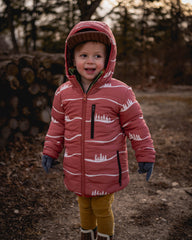 Child wearing a red jacket with white patterns outdoors near stacked logs.