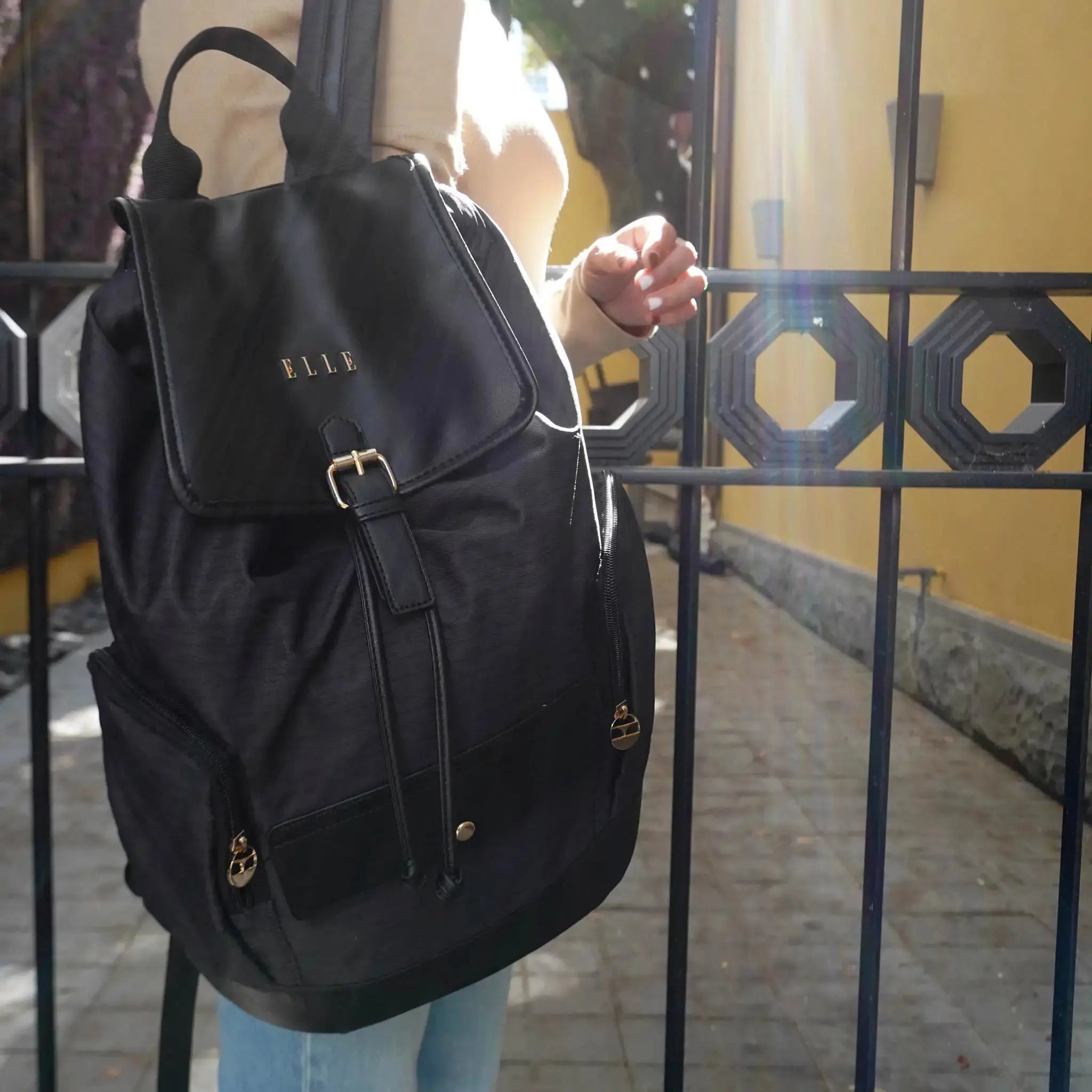Person holding a black backpack with 'ELLE' branding in front of a metal gate.