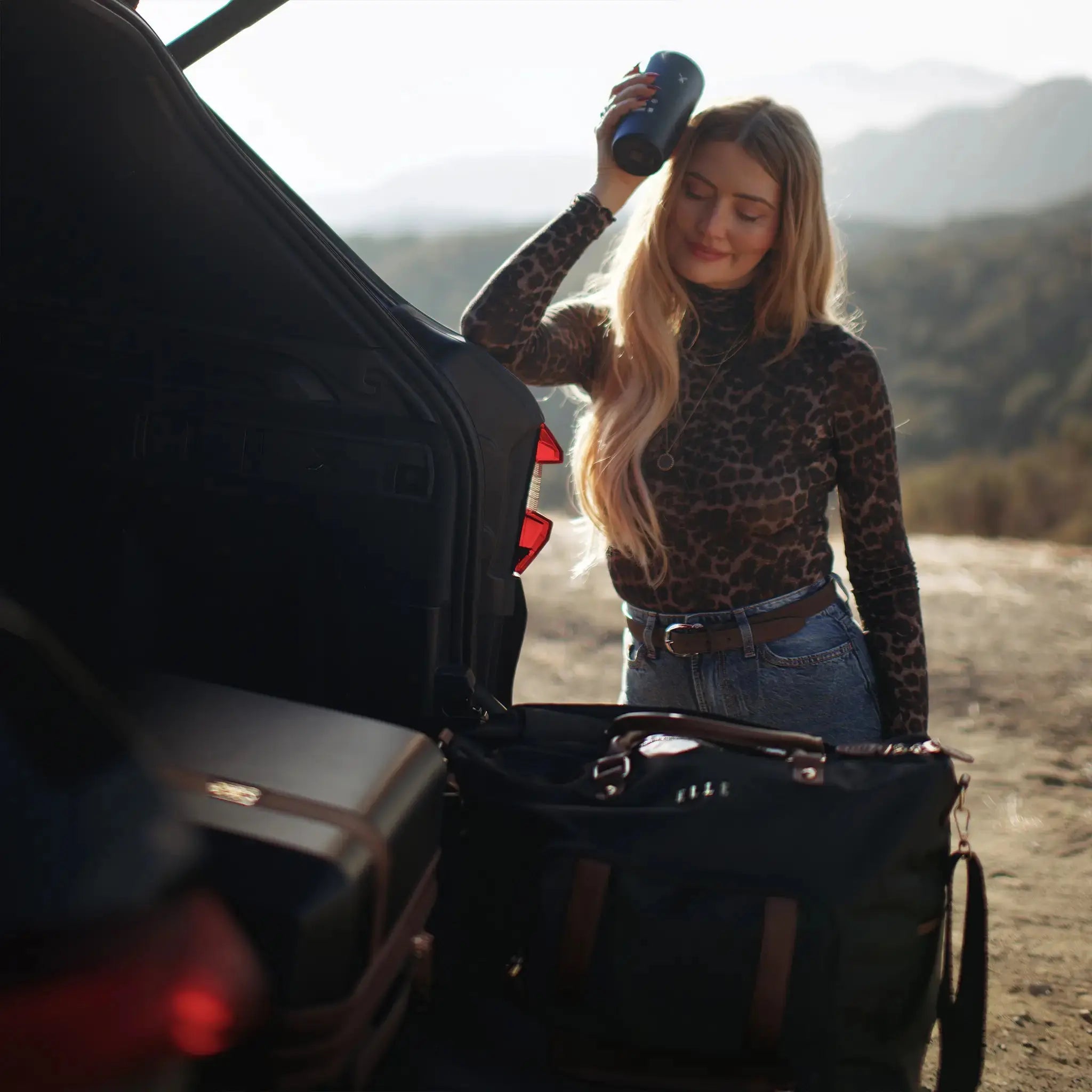 Woman standing next to an open car trunk with a suitcase, mountains in the background