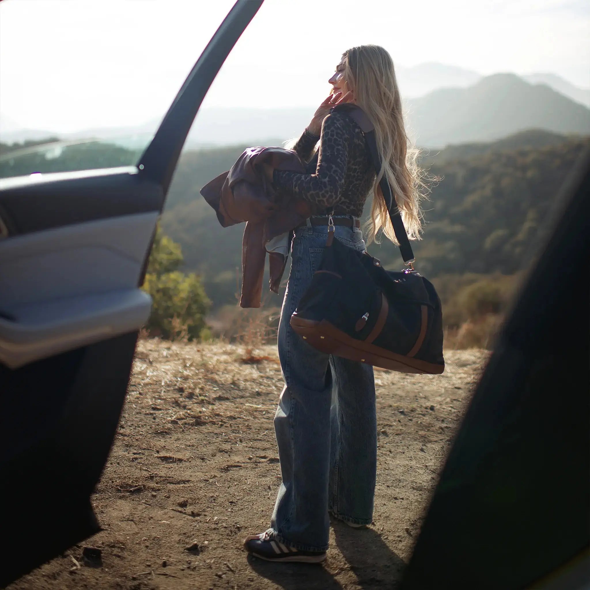 Woman with a bag standing next to an open car door in a scenic outdoor setting