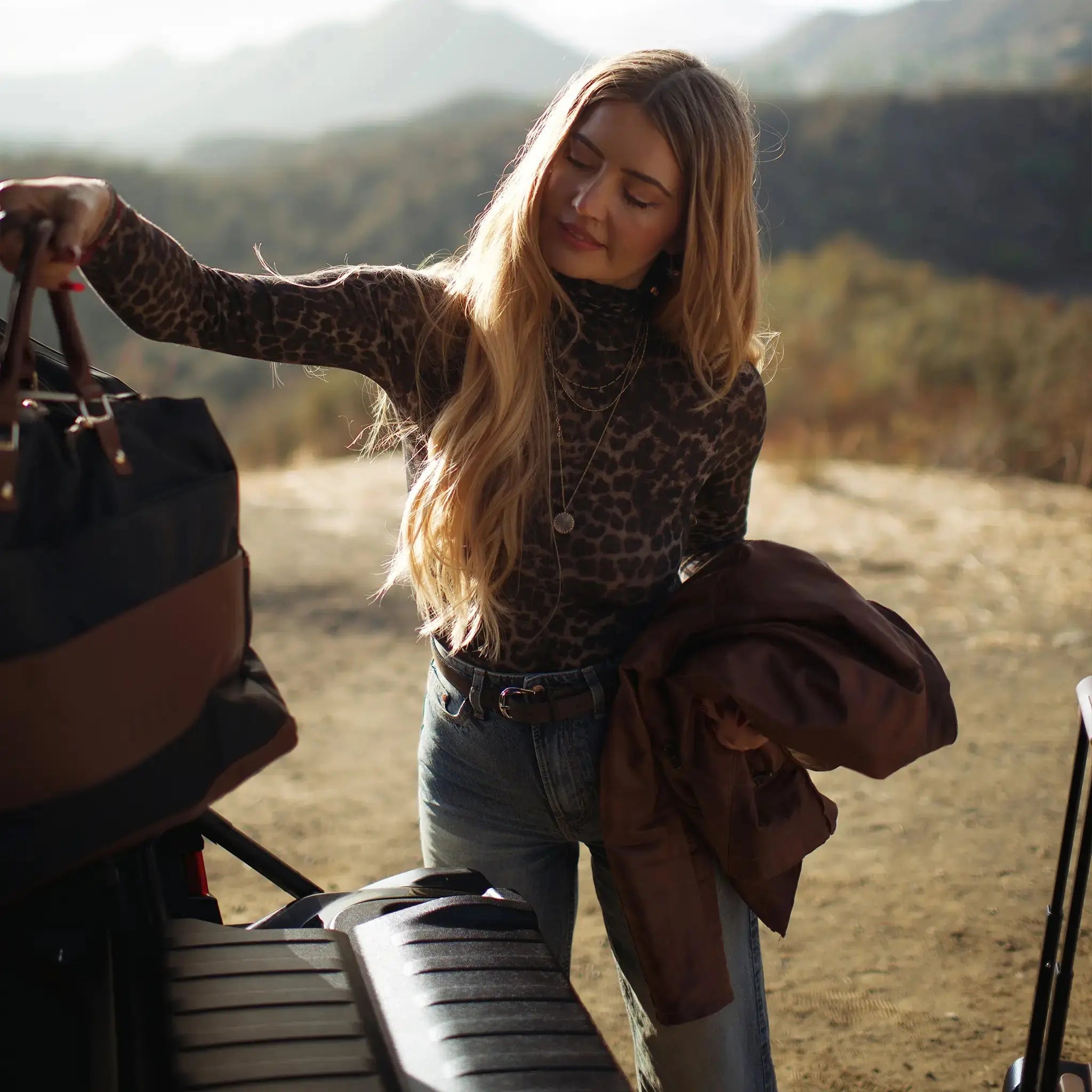 Woman in leopard print sweater and jeans standing next to a vehicle with a suitcase, outdoors.